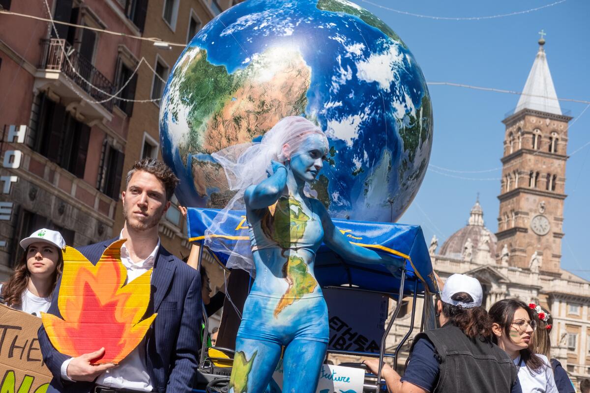 A woman with a partial map of the world emblazoned on her body stands in front of a globe. She is flanked by other people. 
