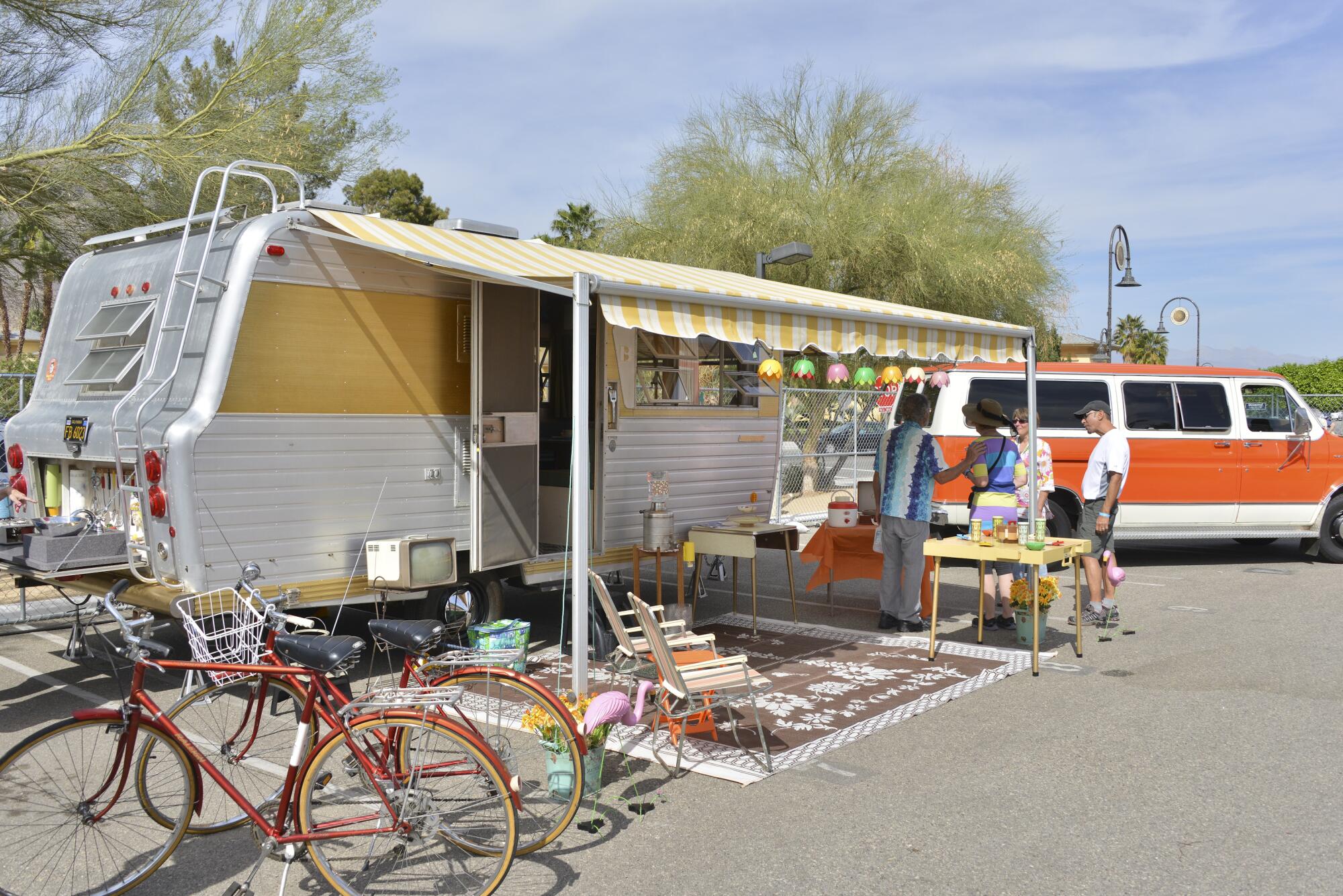 People and bicycles stand outside a parked vintage travel trailer.