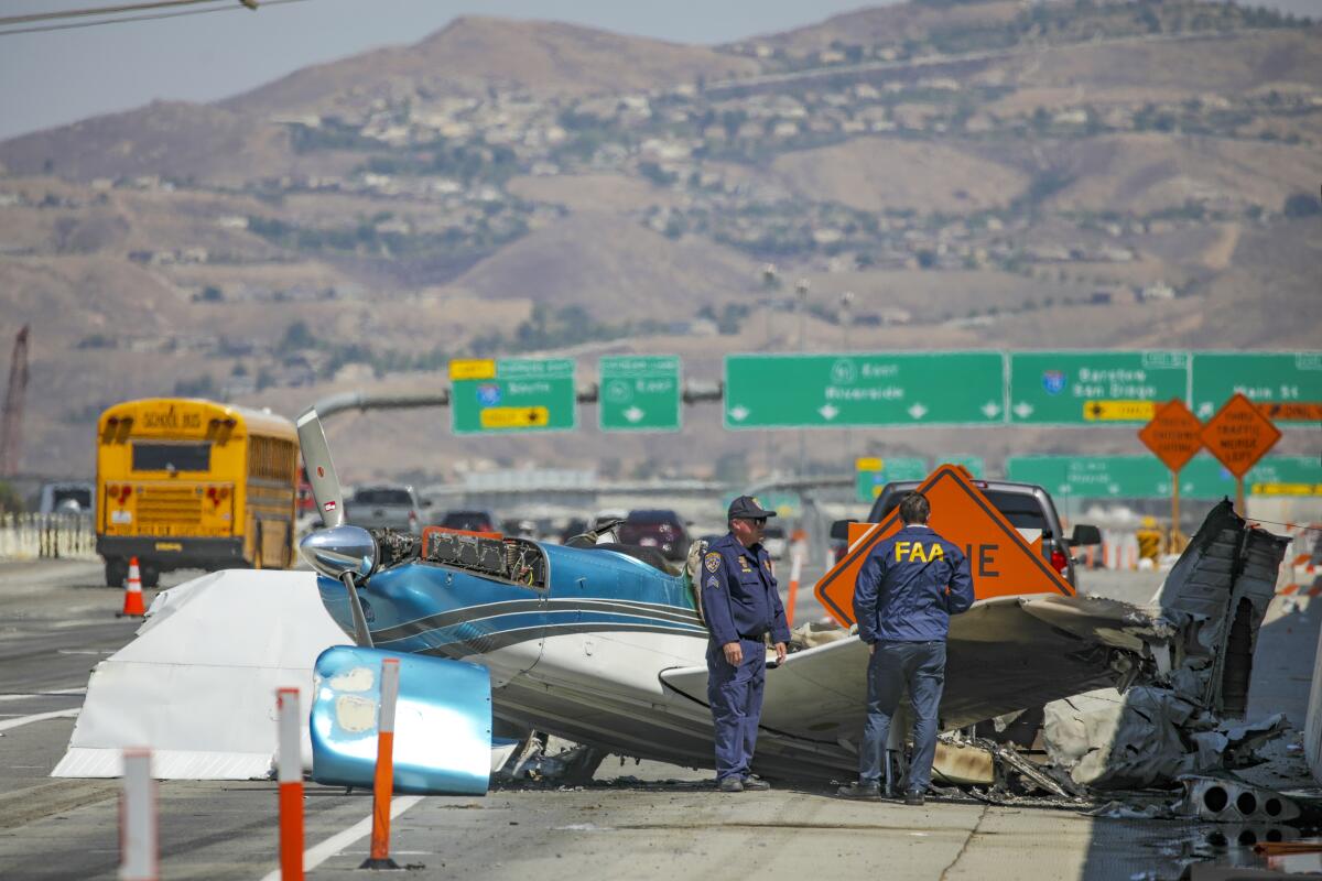 A partially wrecked small plane rests alongside a freeway. Freeway signs are seen in the background.