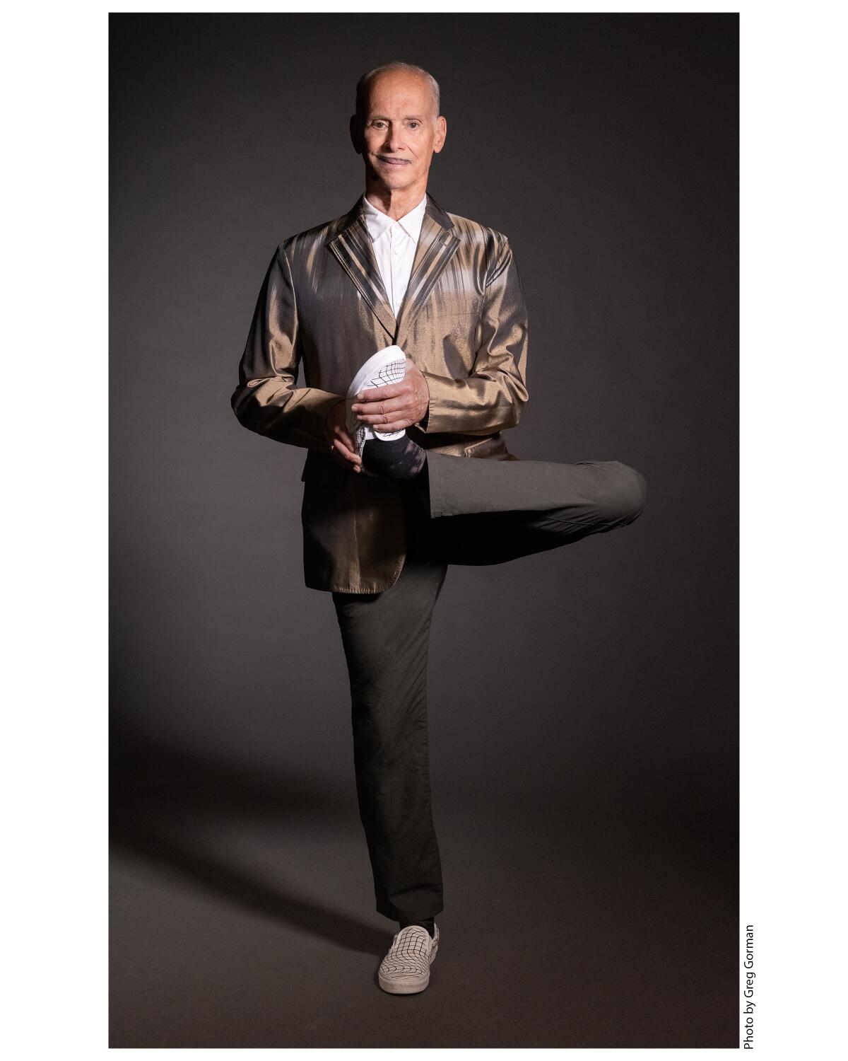 John Waters stands on one leg while holding his other foot. 