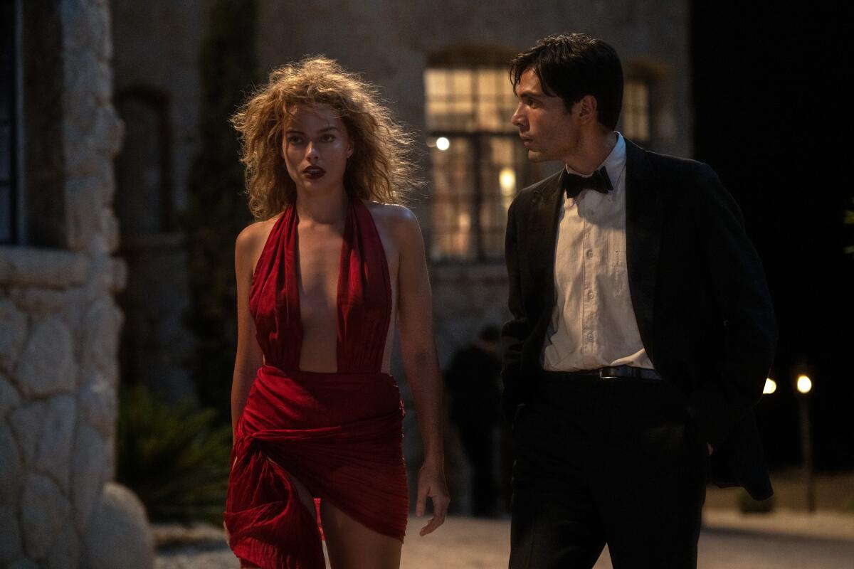 A woman in a red low-cut dress walks outside with a man in a suit in a scene from "Babylon."
