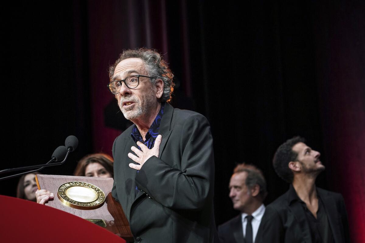 A man with gray hair and glasses holding an award and pressing a hand to his chest while standing in front of a microphone