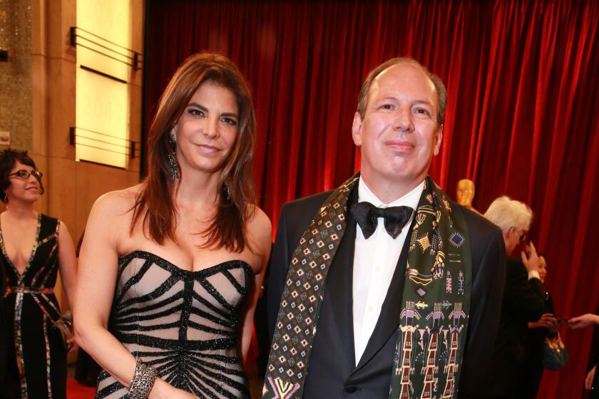 Dina De Luca is wearing a black, sheer, strapless dress alongside Hans Zimmer who is dressed in a black tuxedo and scarf