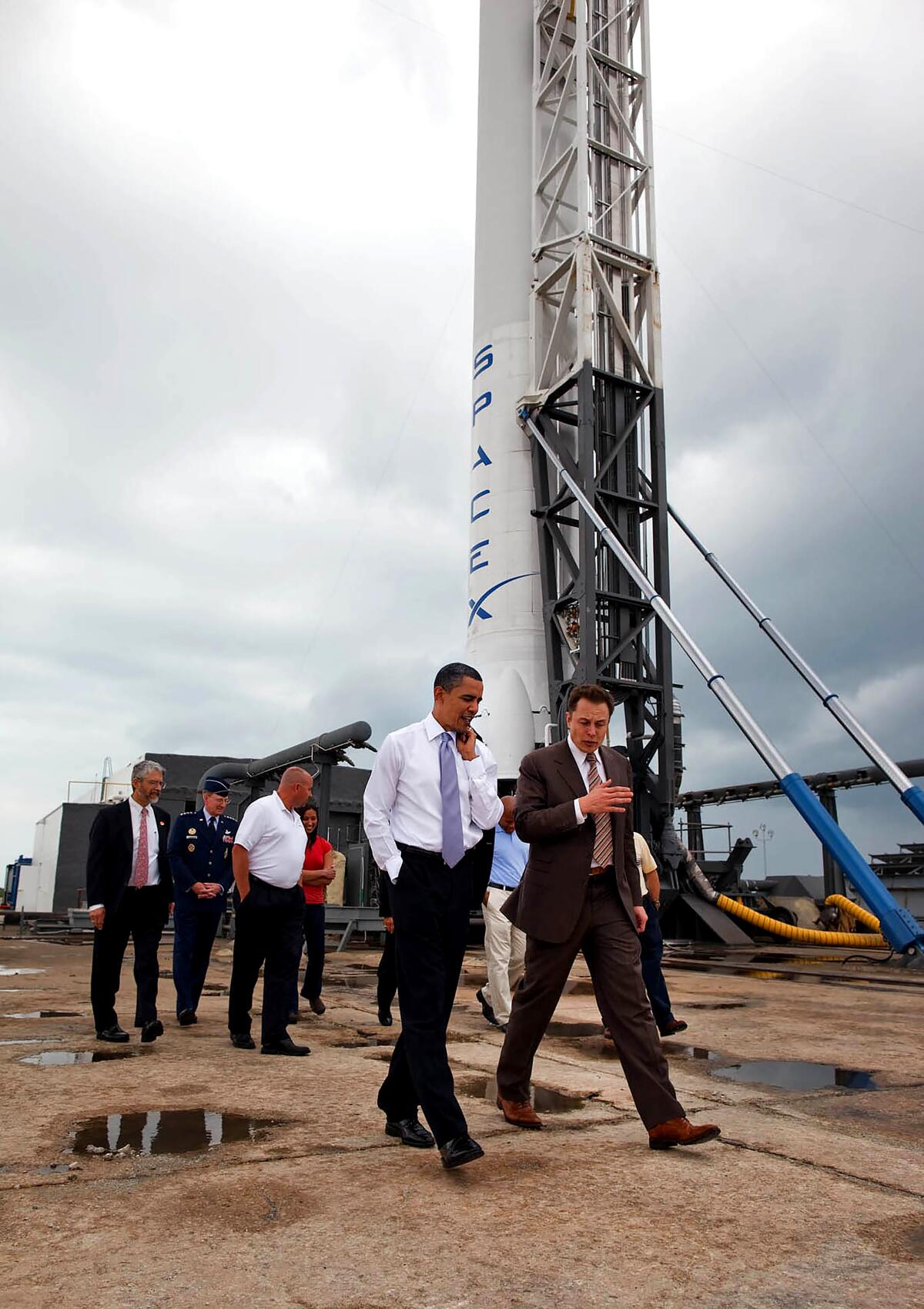 Two men walk past a rocket on the launch pad, with a group of men behind them.