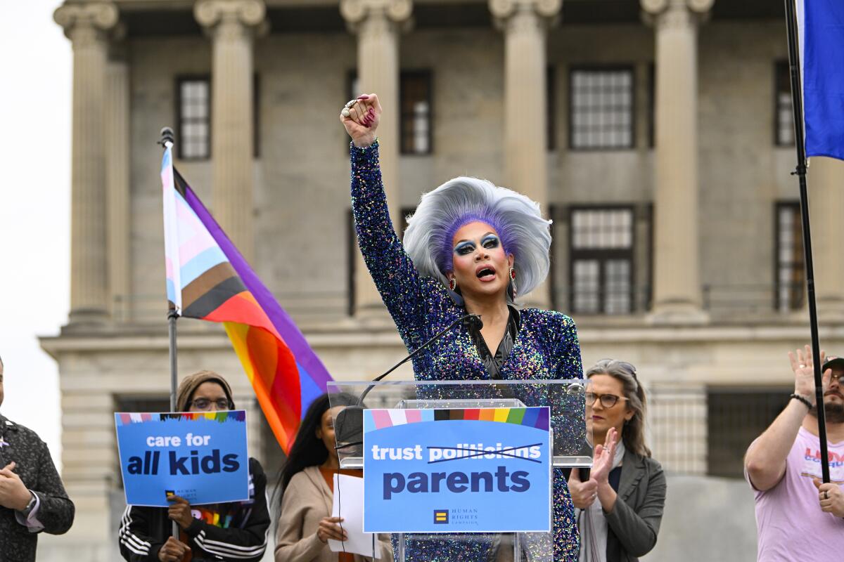 A drag artist raises a fist at a lectern surrounded by people with signs and pride flags