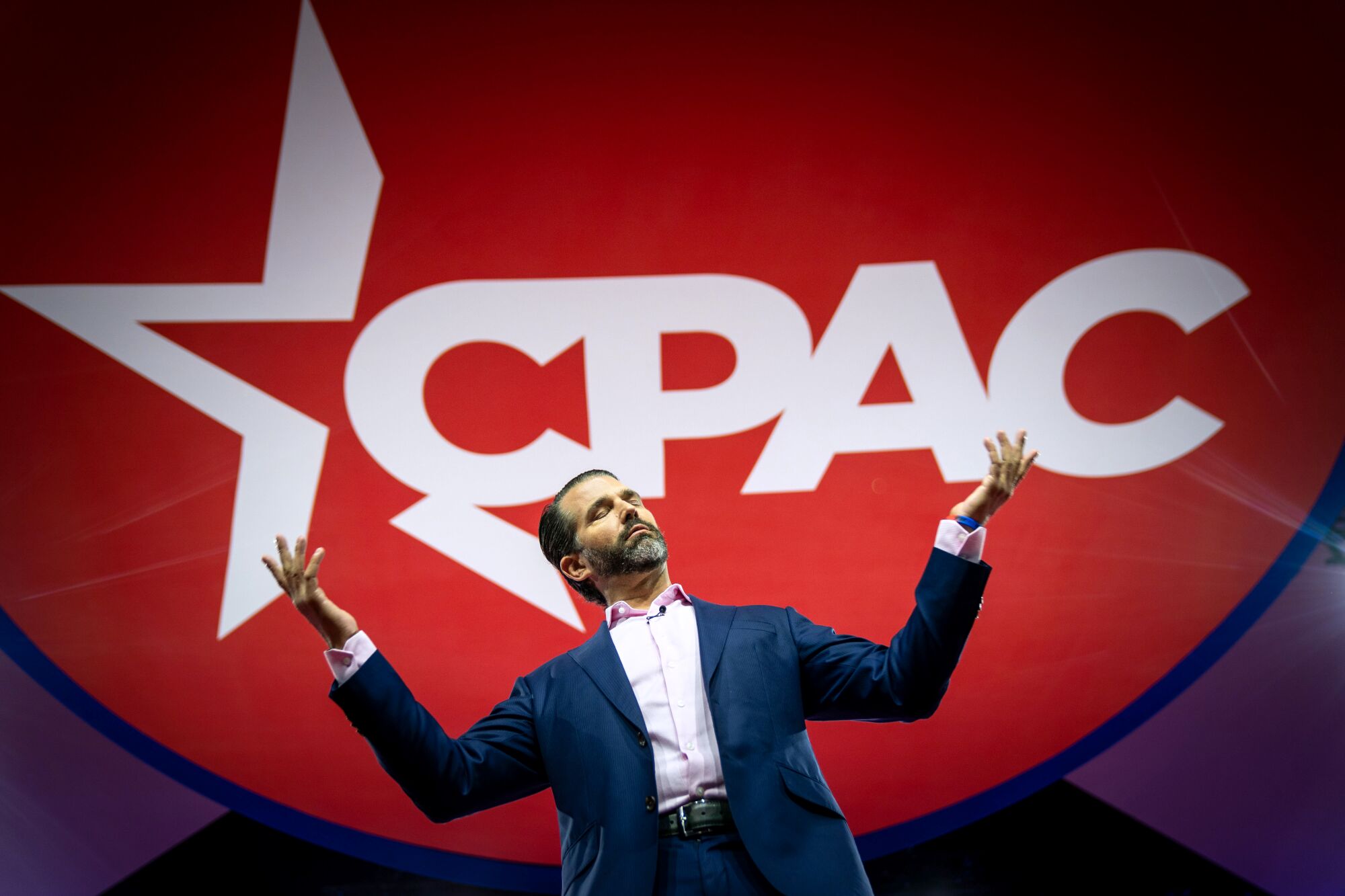 Donald Trump Jr., speaks on stage while white-on-red sign CPAC is displayed behind him.