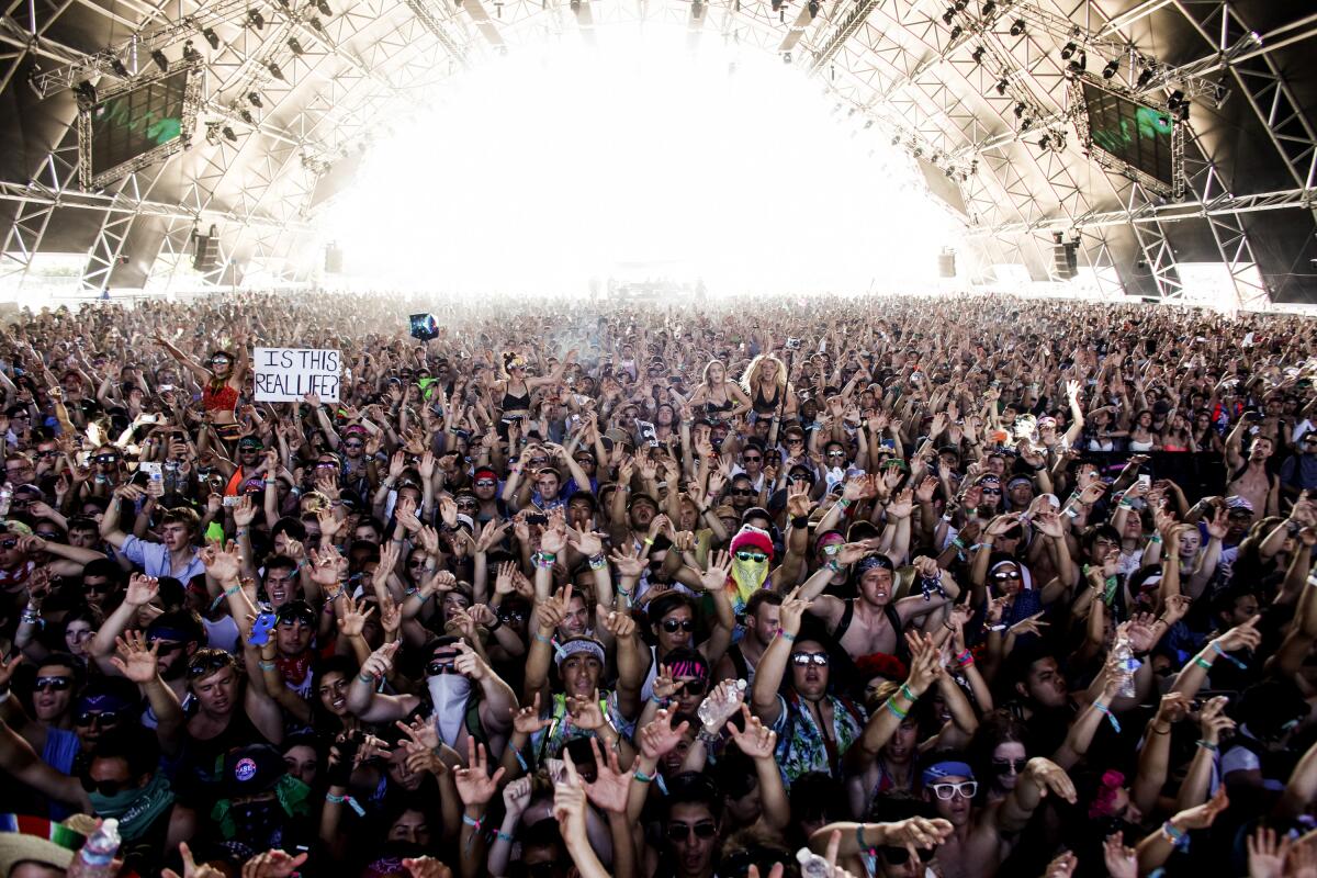 Thousands of concertgoers raise their arms up at an outdoor venue under curved rafters at Coachella.
