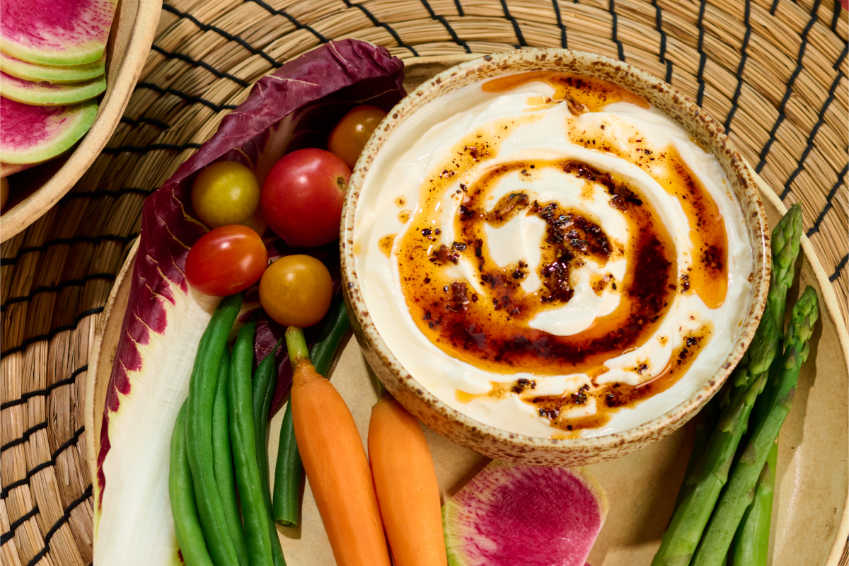 A bowl of white dip with a red chile oil swirl, on a plate with vegetables for dipping