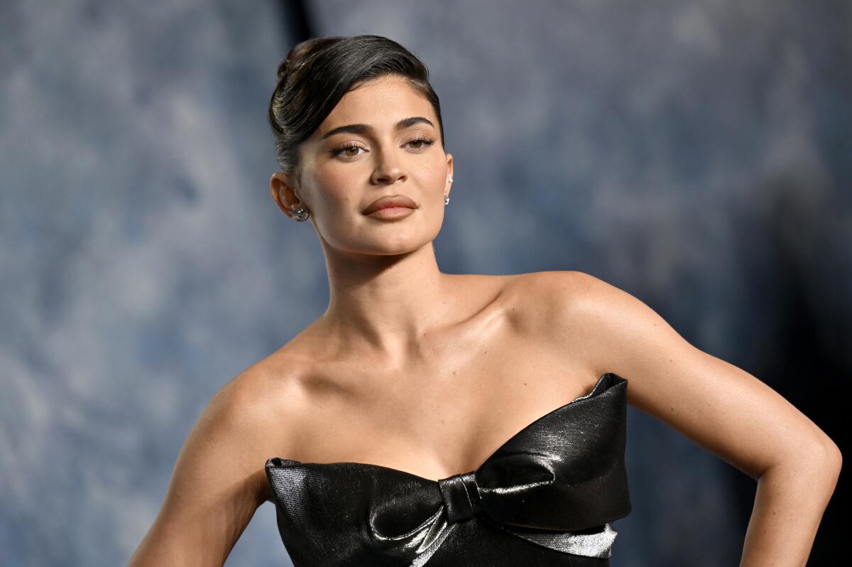 Kylie Jenner poses in front of a gray background with an updo and a shiny black, strapless dress.