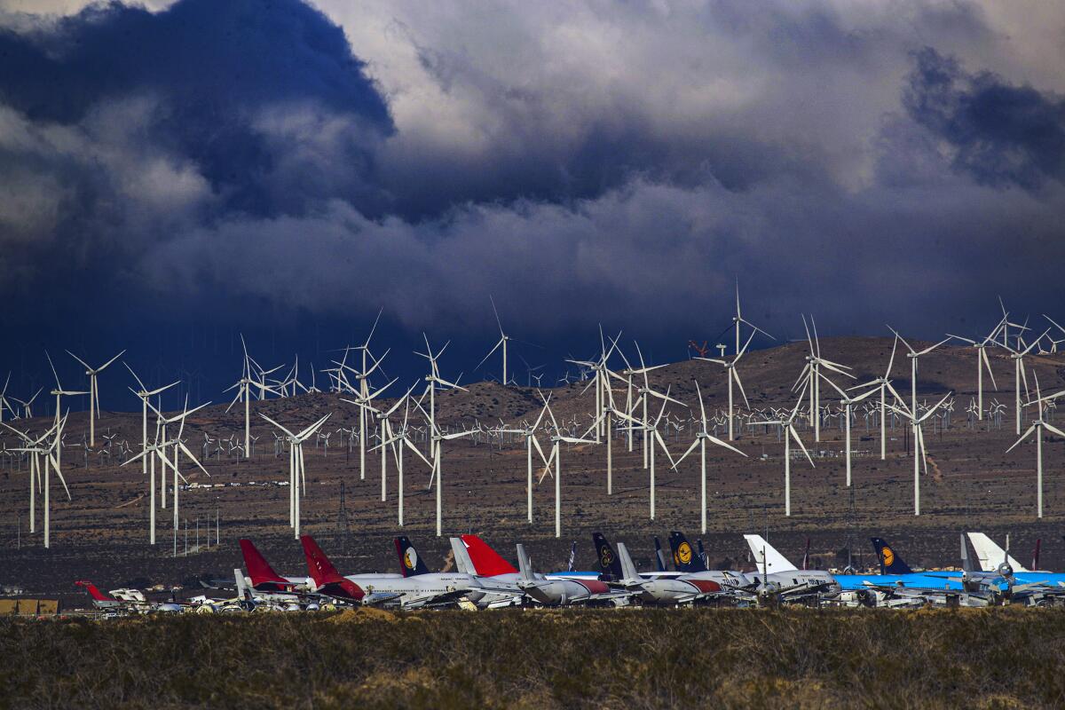 Rows of white wind turbines rise in a desert landscape under stormy skies, with a row of planes in the foreground