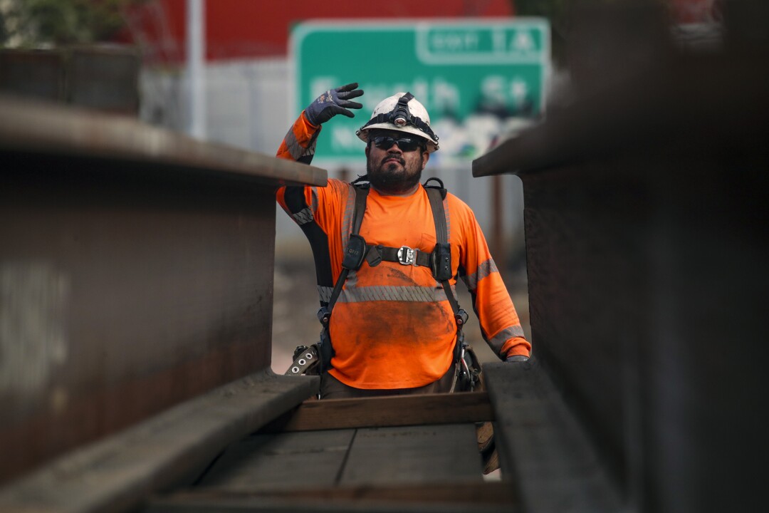 A crew member in helmet and orange shirt gestures with his gloved hand behind beams