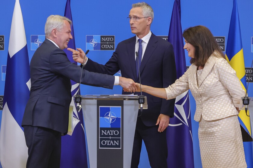 A man and woman shaking hands in front of a man at a lectern with a NATO logo, with flags and NATO logos in the background