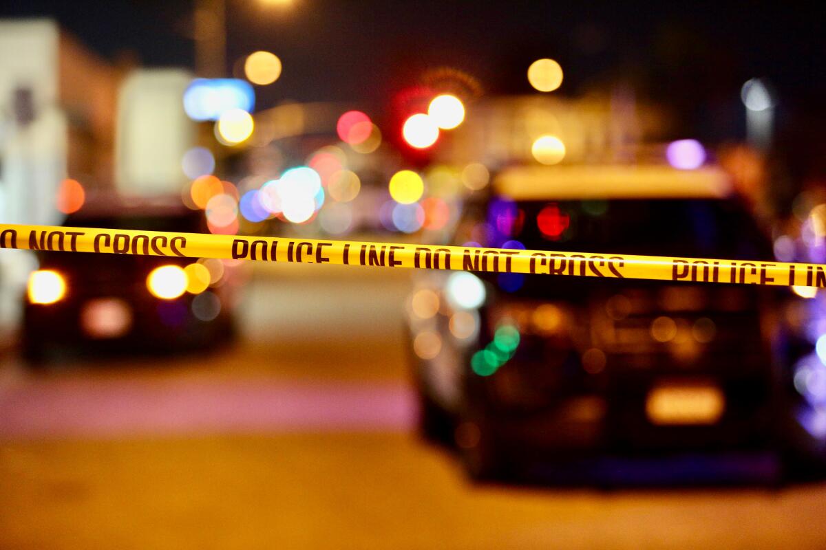 Police tape is strung across a road, with police vehicles seen behind it. 