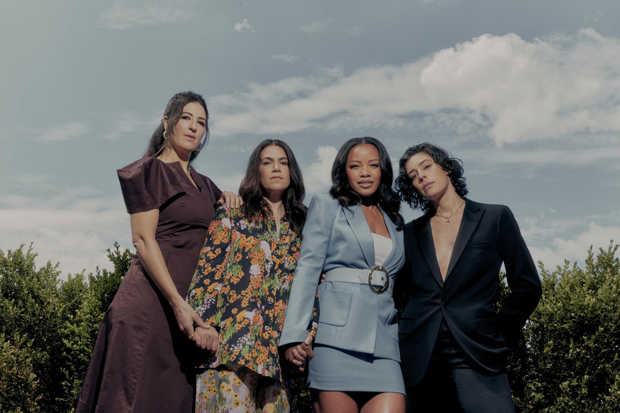 Four women in formalwear pose under a blue sky with white puffy clouds