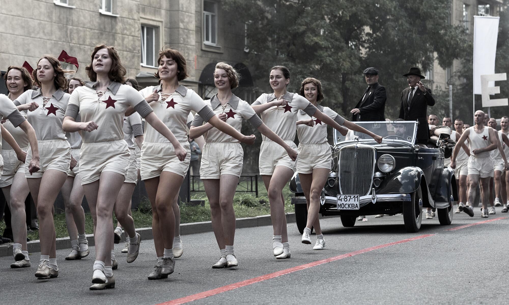 Women in white athletic outfits march in front of a car and marching men.