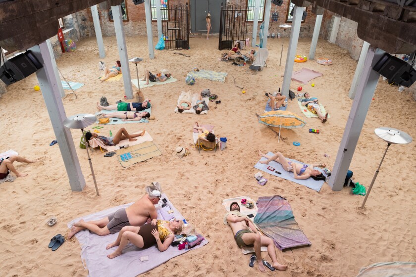 In “Sun & Sea” at the 2019 Venice Biennale, people lie on blankets on sand in a room whose roof is held aloft by columns.