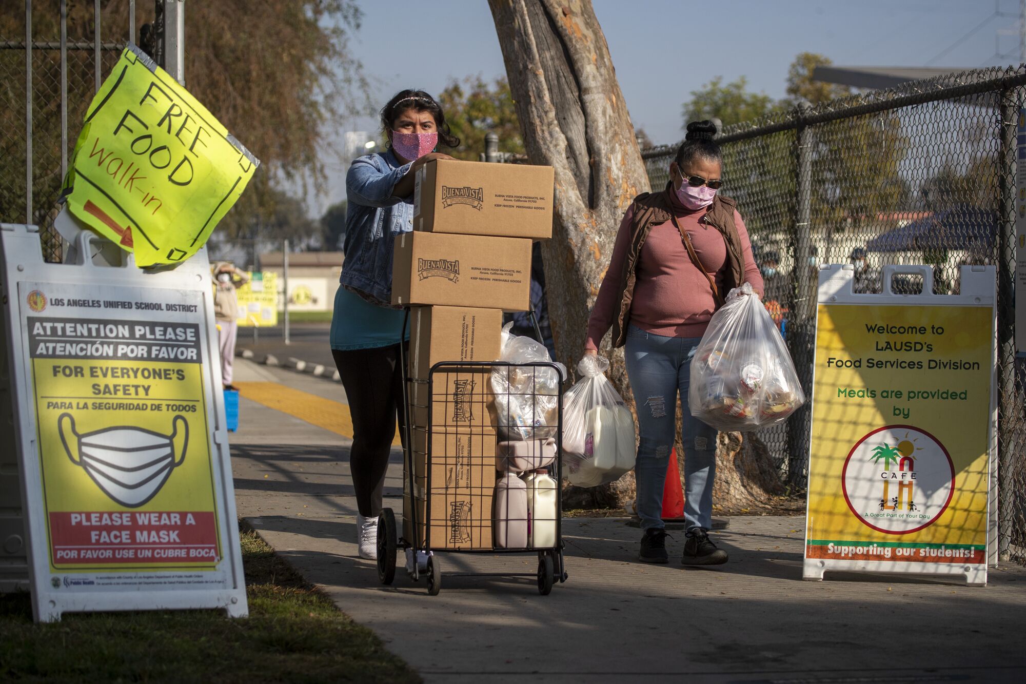 Two women push a cart filled with boxes and carry plastic bags full of food