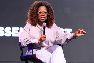 Oprah Winfrey is sitting on stage talking into a microphone and gesturing with her hand while wearing glasses