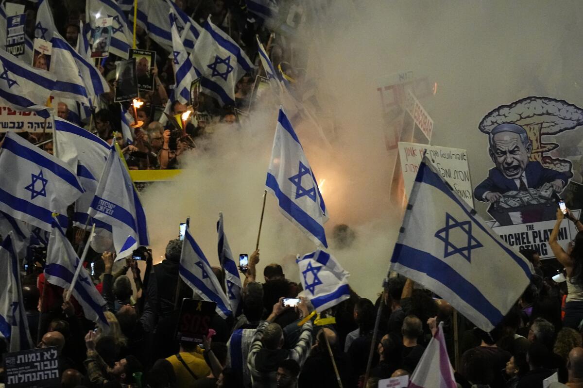 Protesters in Tel Aviv hold Israel flags and signs mocking Prime Minister Benjamin Netanyahu as smoke rises from the ground.