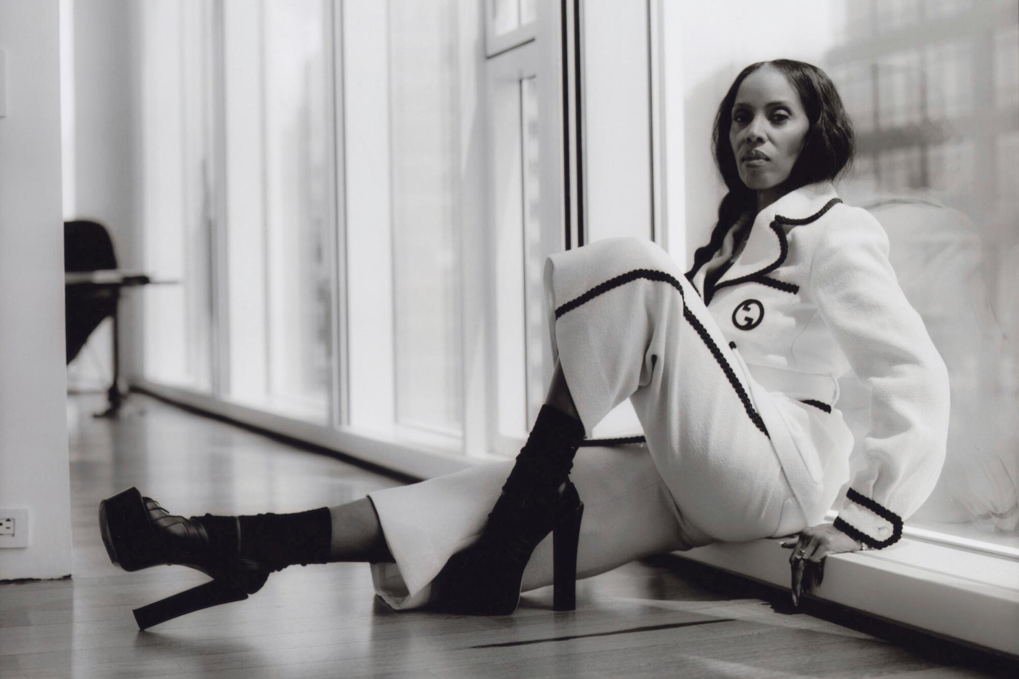 June Ambrose, sitting on the floor of an apartment, wears a white suit and black boots.