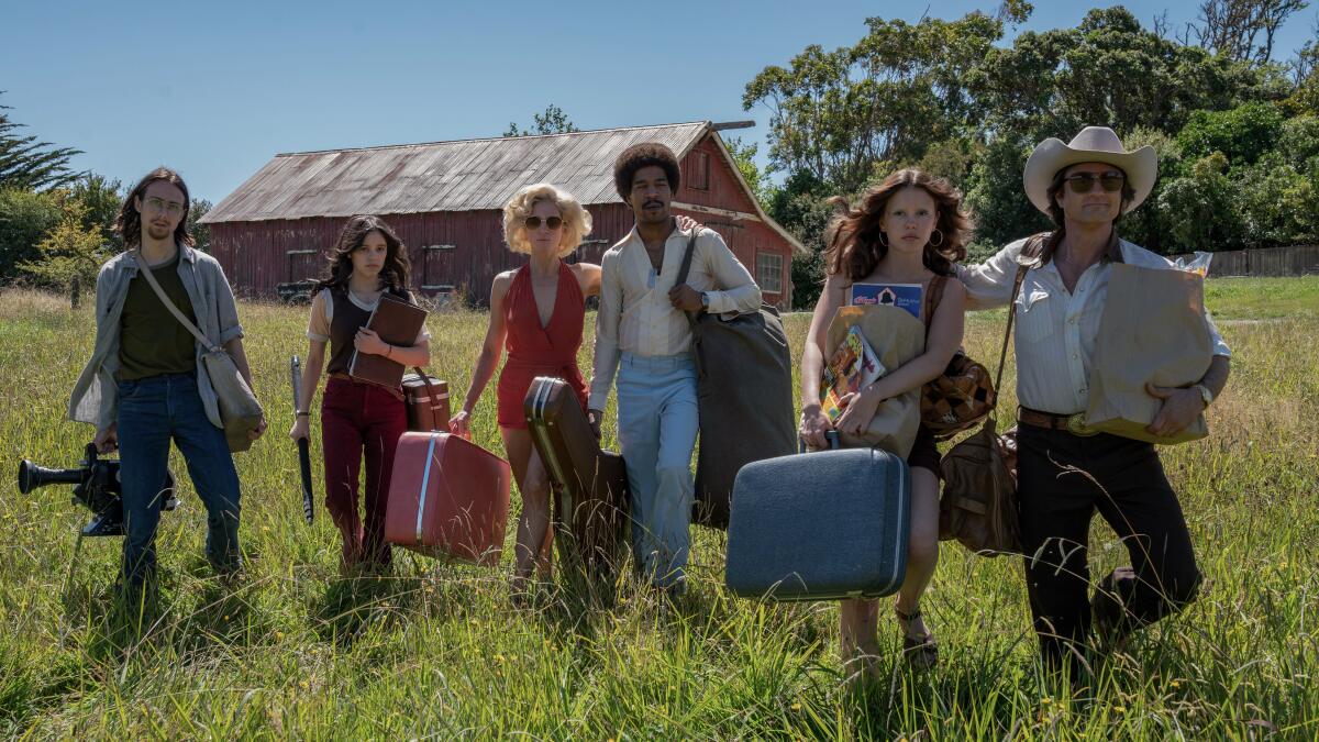 A group of people carrying luggage through a field, with a barn in the background.