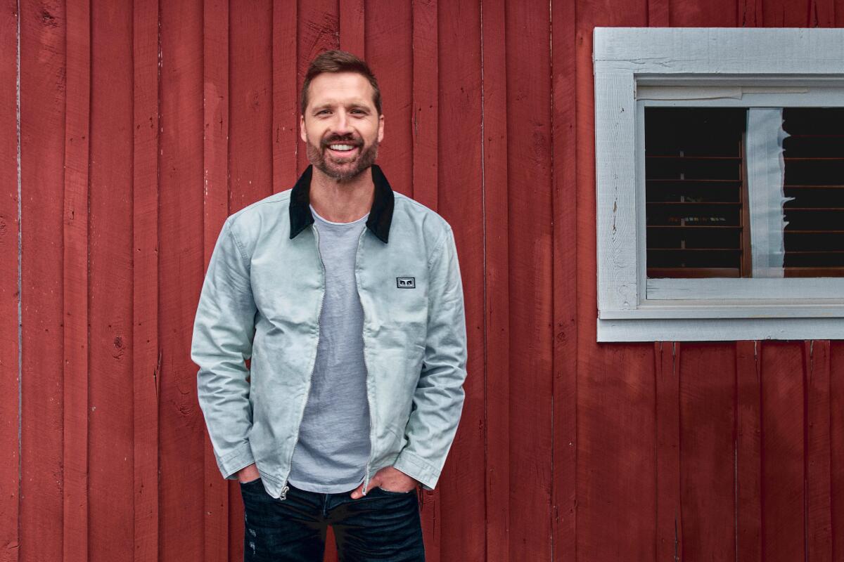 A man in a gray jacket stands outside a red barn 