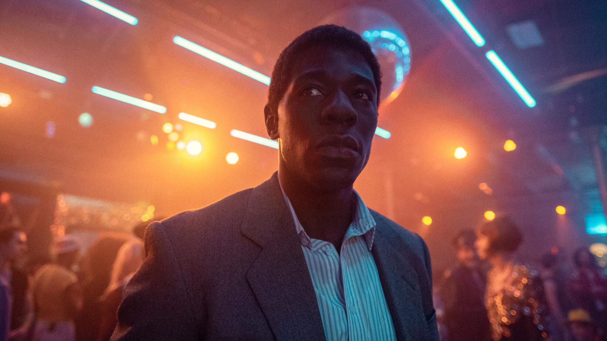A man in a shirt and jacket stands in a nightclub.