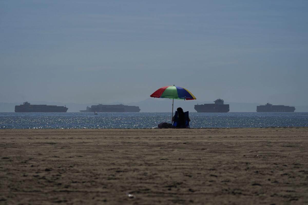A person lounges on a beach in Seal Beach as container ships wait to dock.