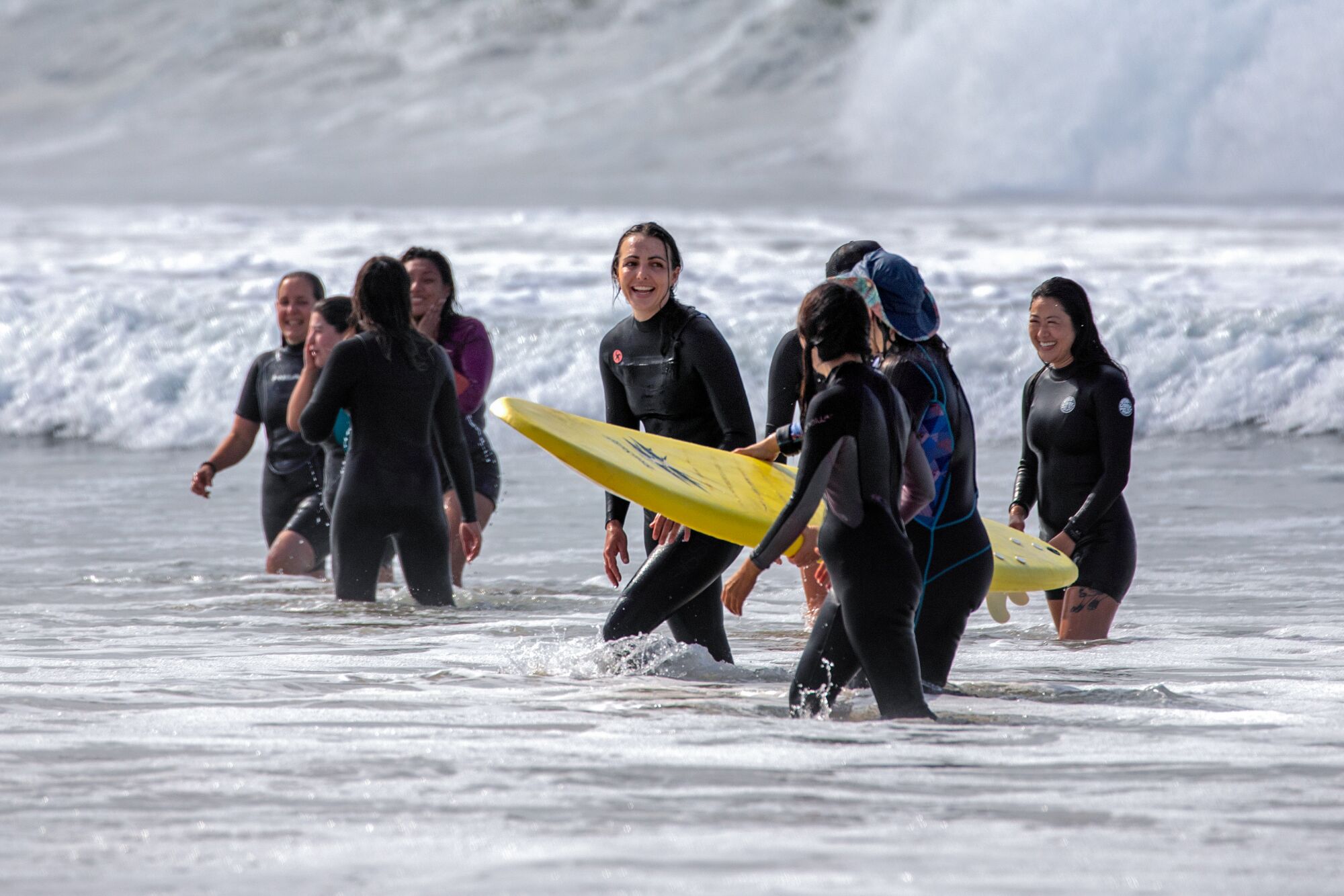 Women in wetsuits walk in shallow ocean water. One carries a yellow surfboard.
