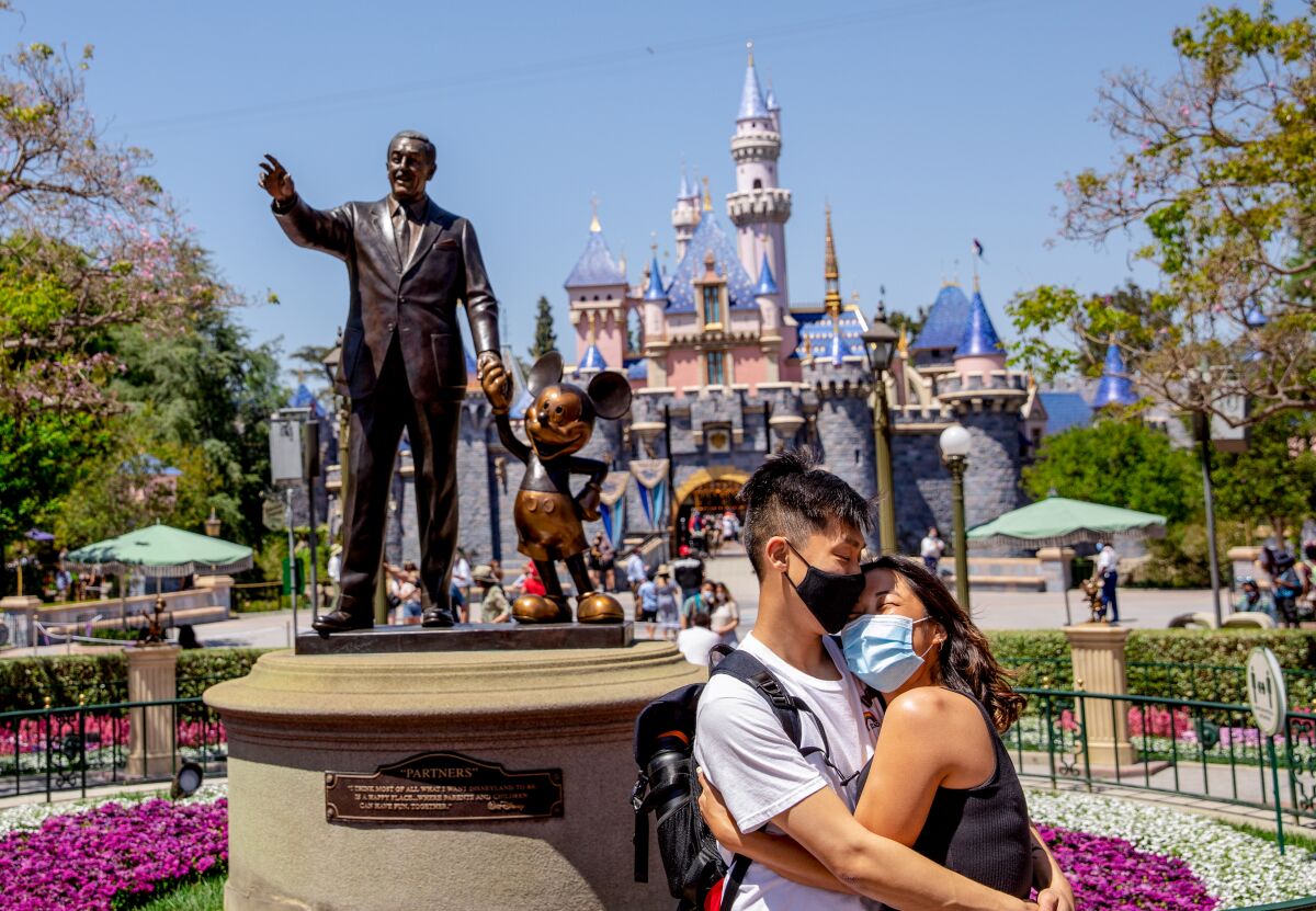 A masked couple embraces in front of a statue of Walt Disney and Mickey Mouse