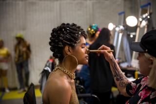 A makeup artist brushes makeup onto a performer's face in a green room behind the scenes of a TV production.