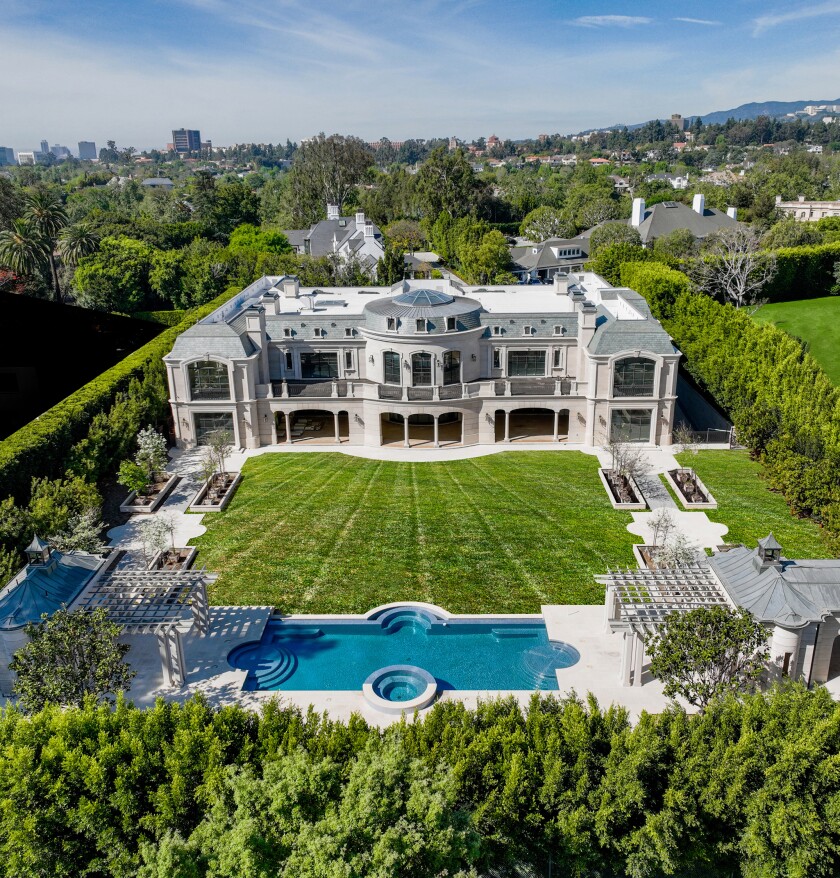 A mansion in Holmby Hills has a pool and lawn in front and is surrounded by tall hedges. 