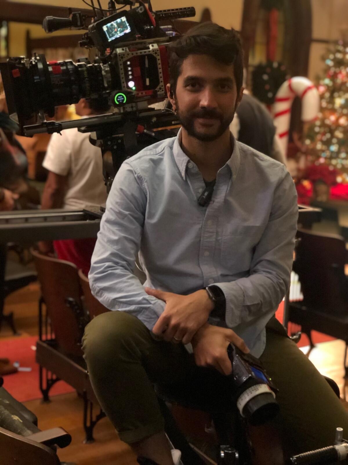 Ian Barbella, in a blue collared shirt and green pants, holds a camera on set.