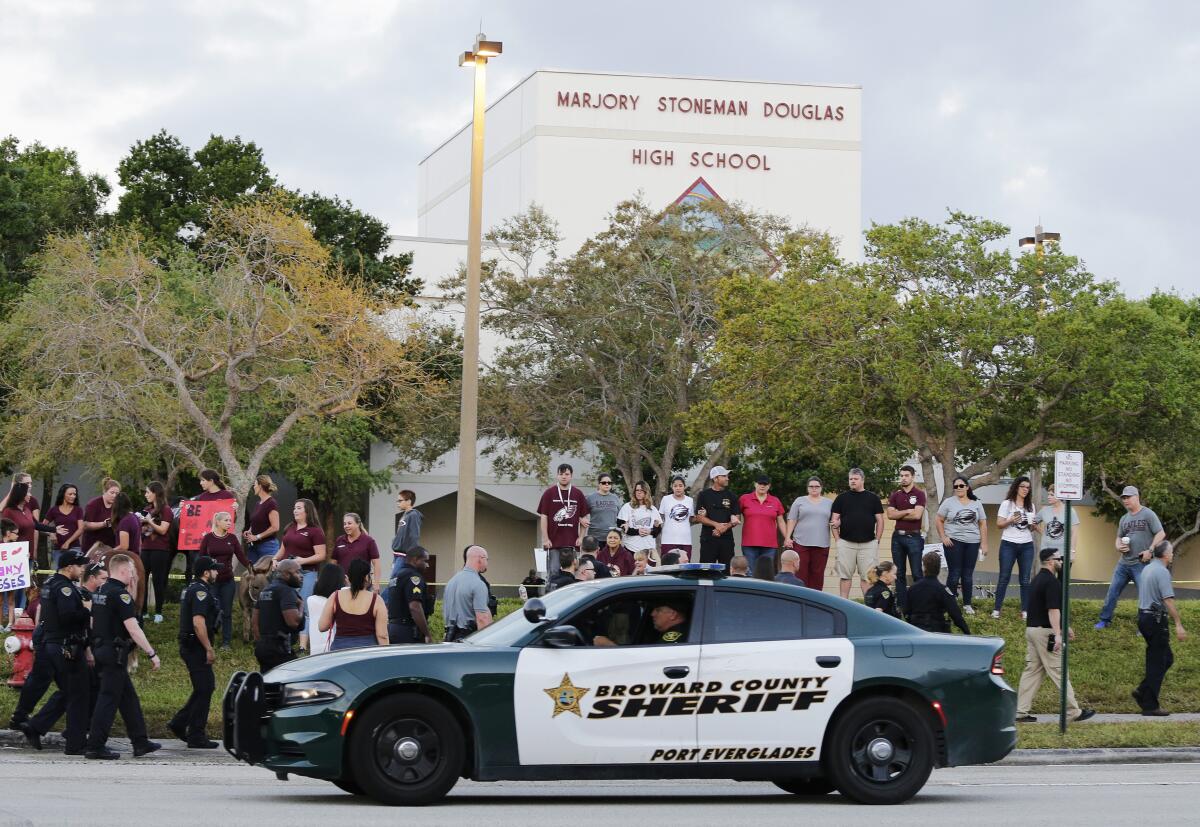 A police car drives by Marjory Stoneman Douglas High School in Parkland, Fla., as people, including police, stand nearby.