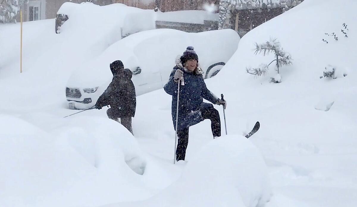 Two people try to navigate through deep snow. A car is buried under snow.