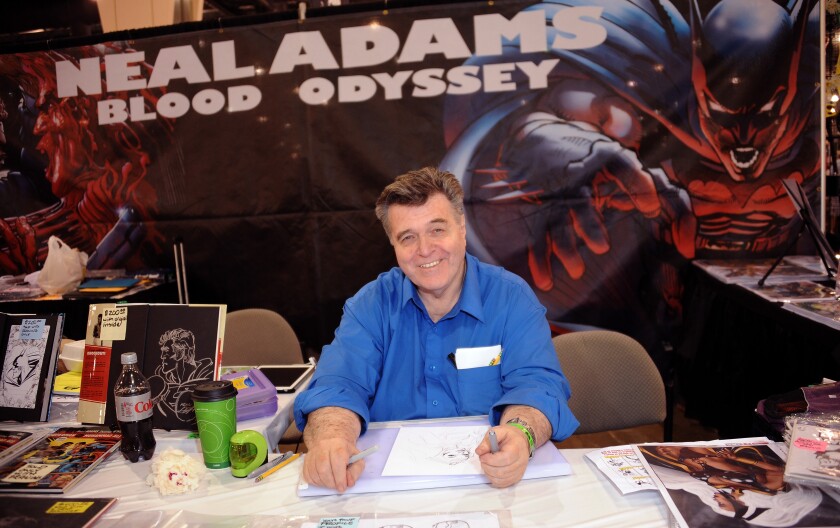 Artist Neal Adams sits behind a desk with pen in hand