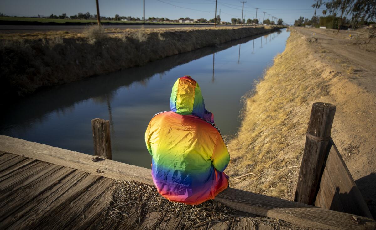 A person in a rainbow-colored hooded jacket sits by a canal.