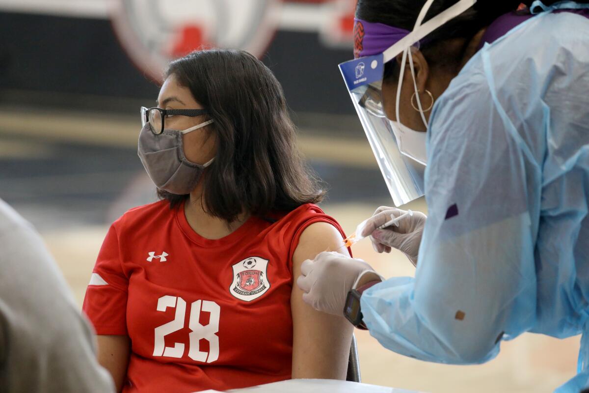 A masked teenager in glasses and a red shirt gets a shot in the arm from a health worker