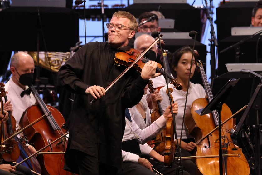 A violinist plays in front of seated orchestra musicians.