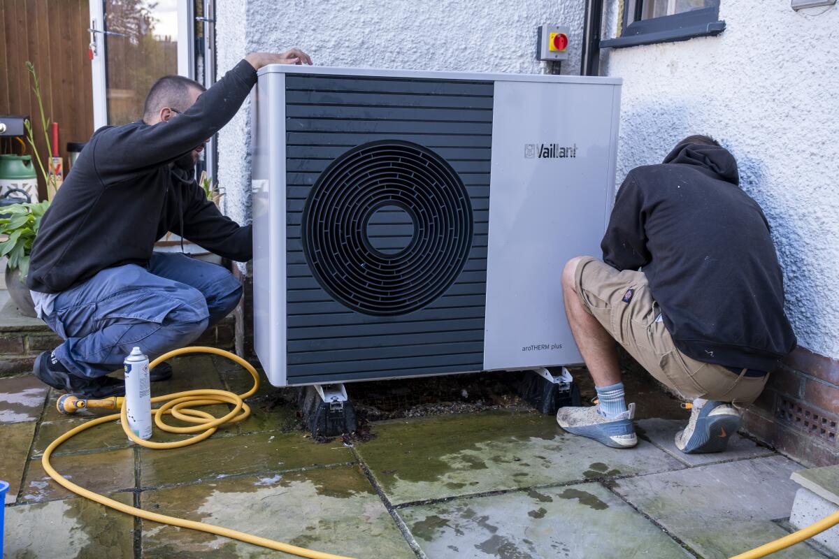 Two men kneel while working on a heat pump.