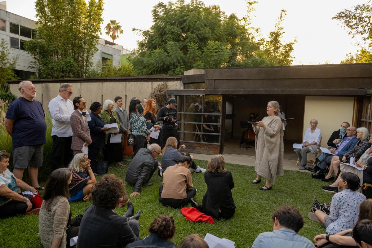 A woman in a loose brown dress performs before an audience seated and standing in a garden.