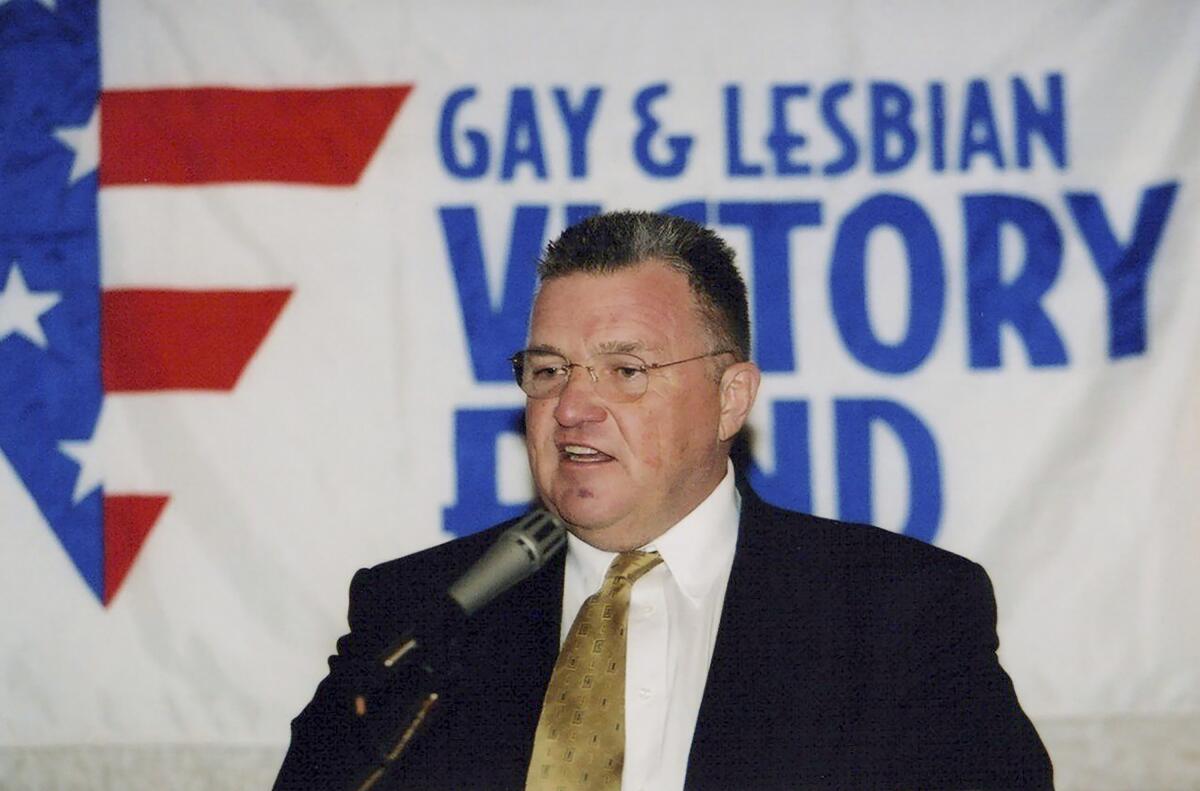 David Mixner, with short hair, glasses and wearing a dark suit and gold tie, speaks into a microphone.