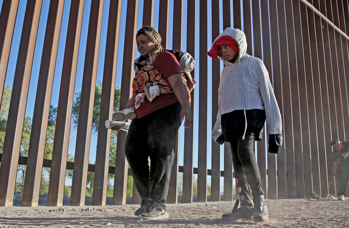 A woman carrying one child and walking alongside another next to wall made of a series of tall posts