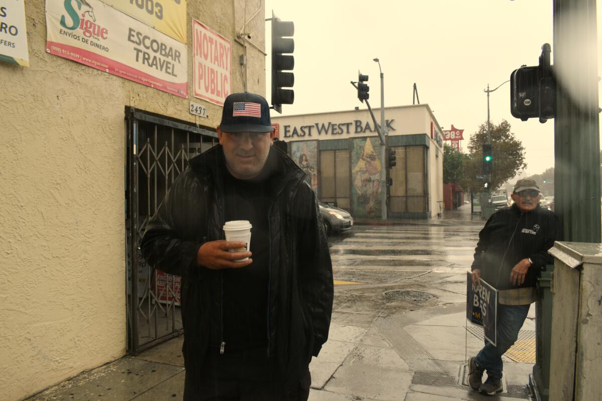 A man in an American flag cap holding a hot drink in the rain, near another man holding a Karen Bass campaign sign.