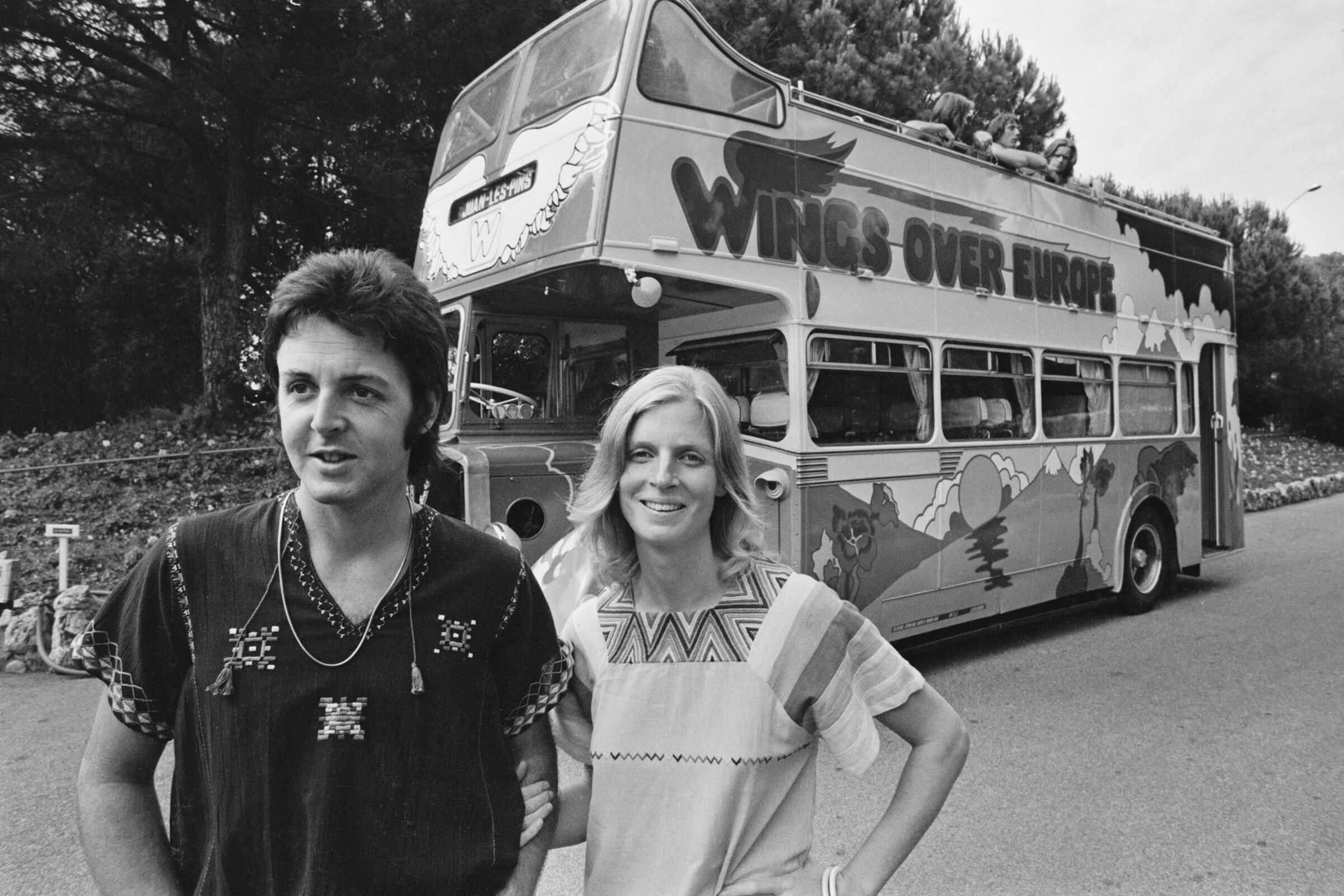 Paul and Linda McCartney, in embroidered shirts, stand in front of a bus that says "Wings over Europe."