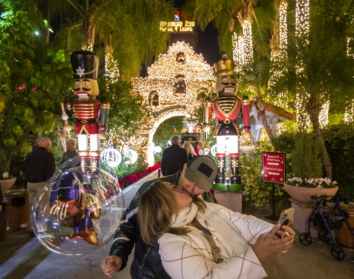 A man and a woman kiss and take photos among the sights and sounds of a holiday light display.