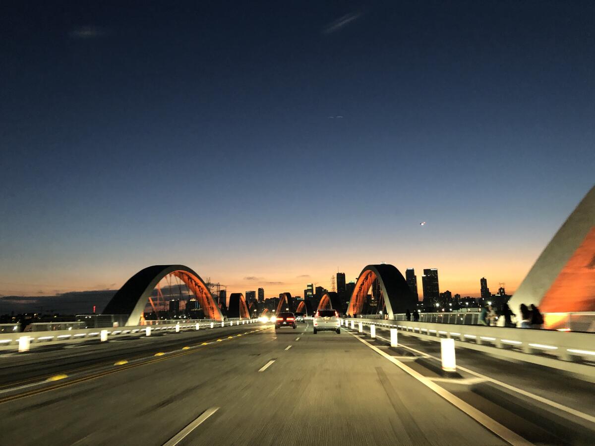 The 6th Street Bridge's arches are bathed in red light at dusk, with the outline of downtown L.A. visible in the distance.