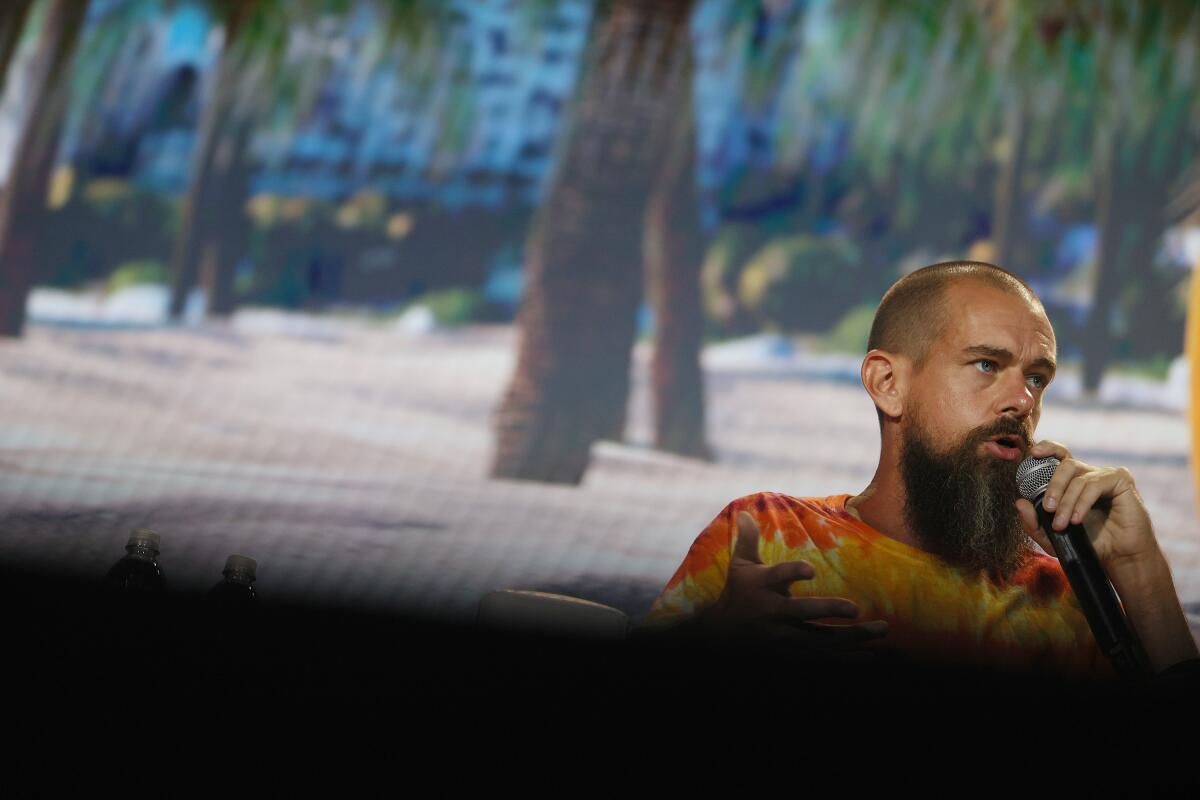 A man in a multi-colored tie-dye shirt with a shaved head and six-inch beard speaks into a microphone.