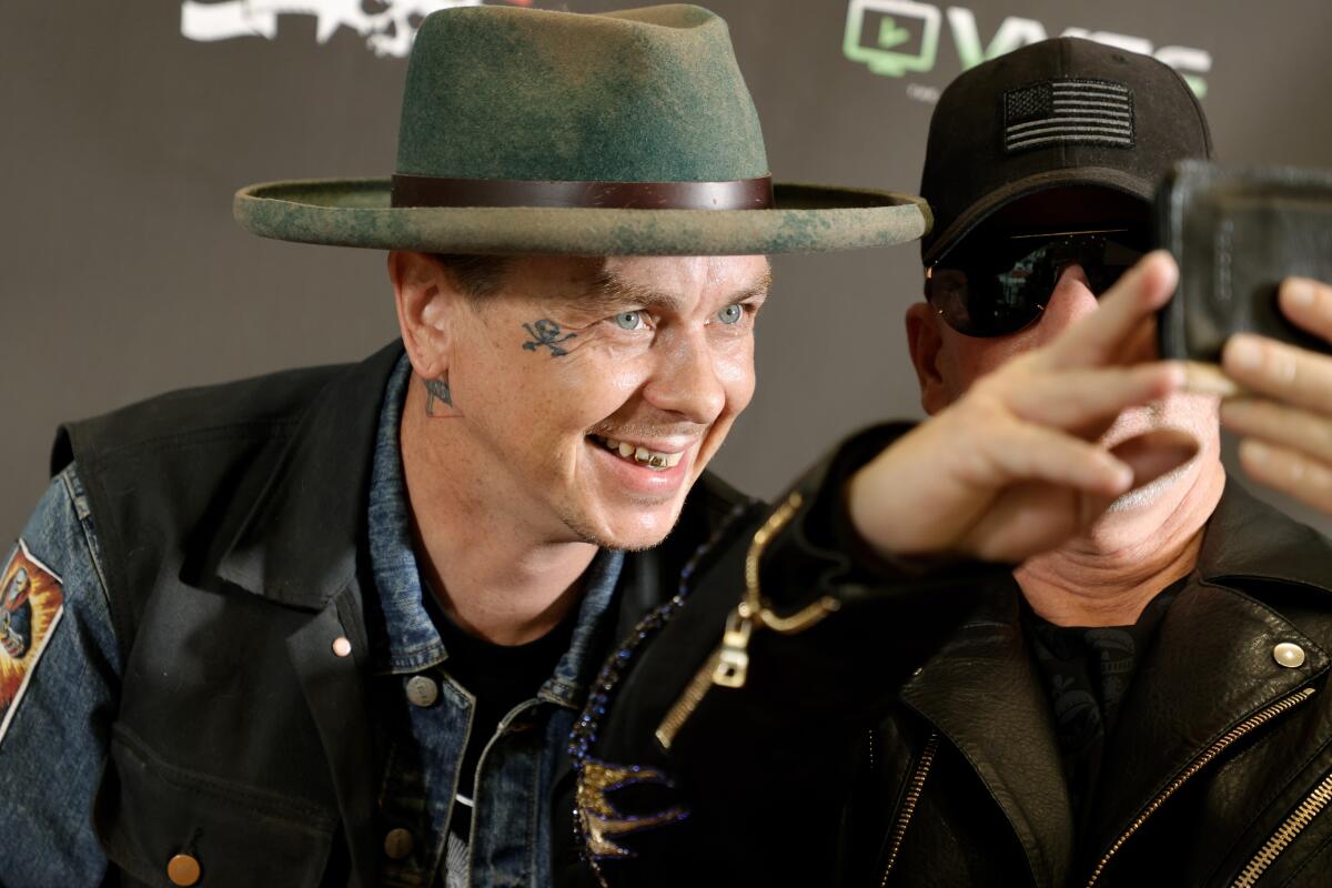 Sid Wilson in a large hat leaning over and smiling while next to a man taking a selfie