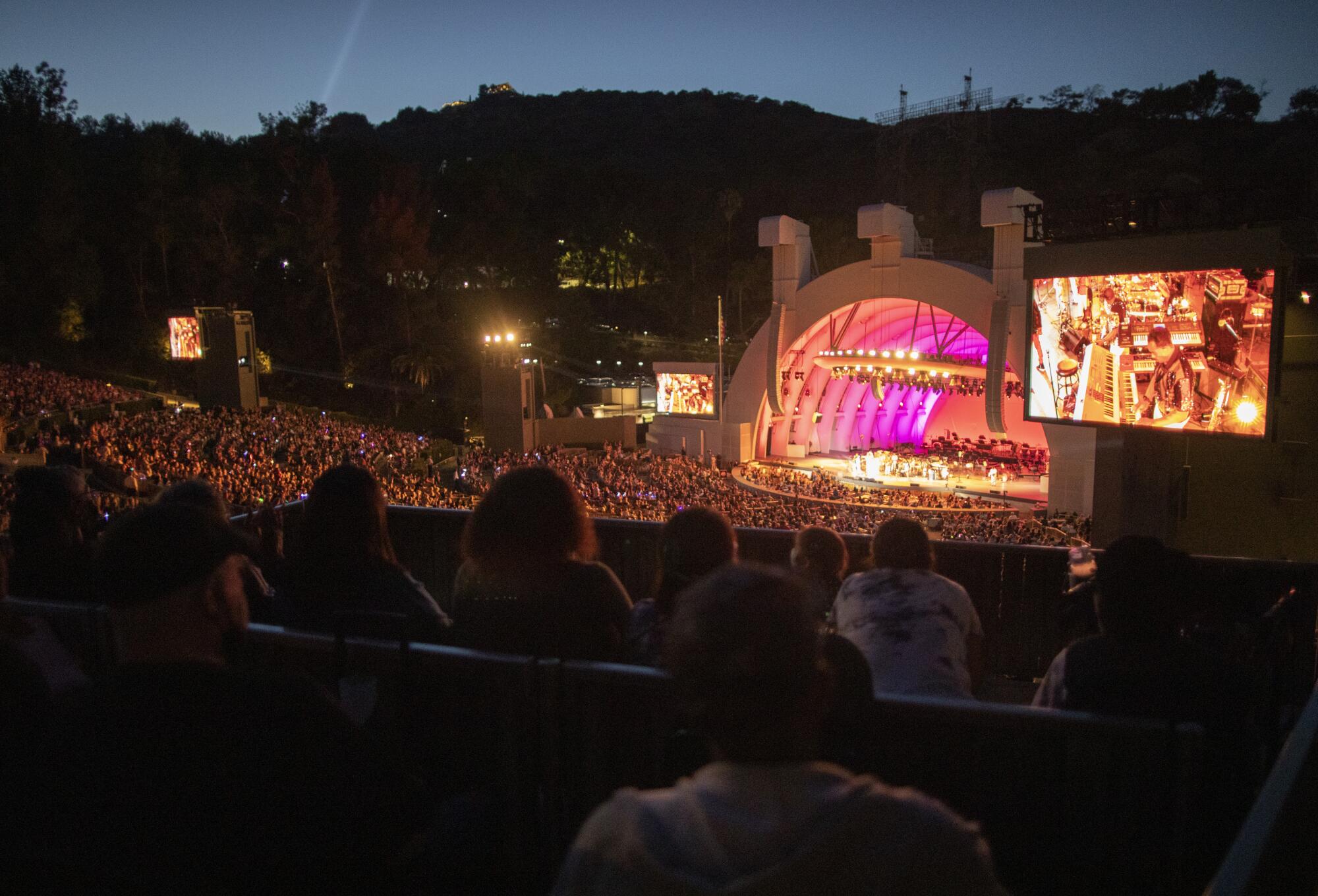 The packed audience watches a big screen and the orchestra on stage, which glows with lighting, at dusk.
